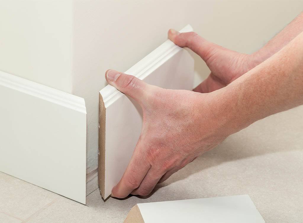 A person installs a piece of white trim on the wall as part of skirting repair work.